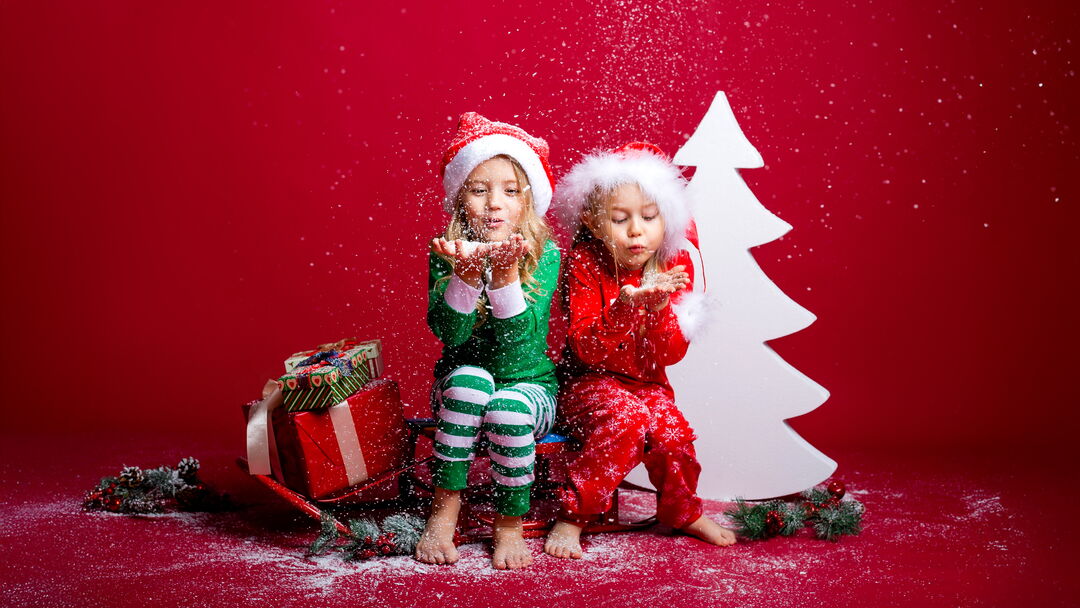 Zwei Kinder in Weihnachtspyjamas und Nikolausmützen auf Schlitten mit Geschenken pusten Schneeflocken