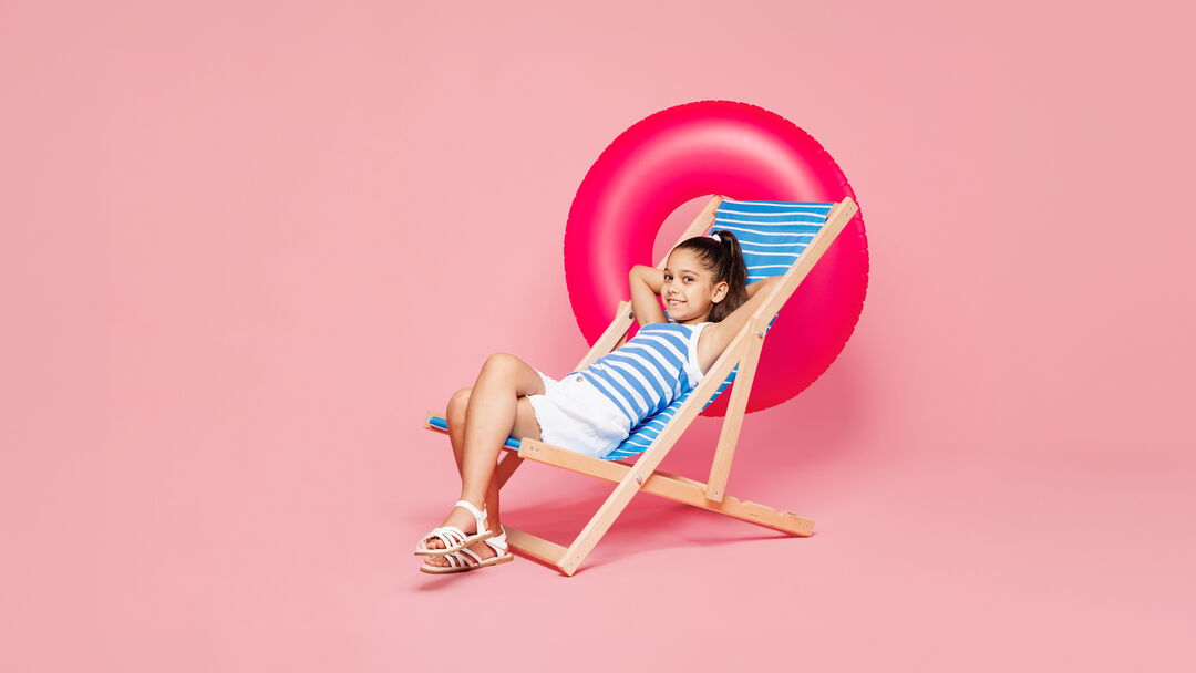 Mädchen in Sommerkleidung auf Liegestuhl mit pinkem Schwimmring im Hintergrund